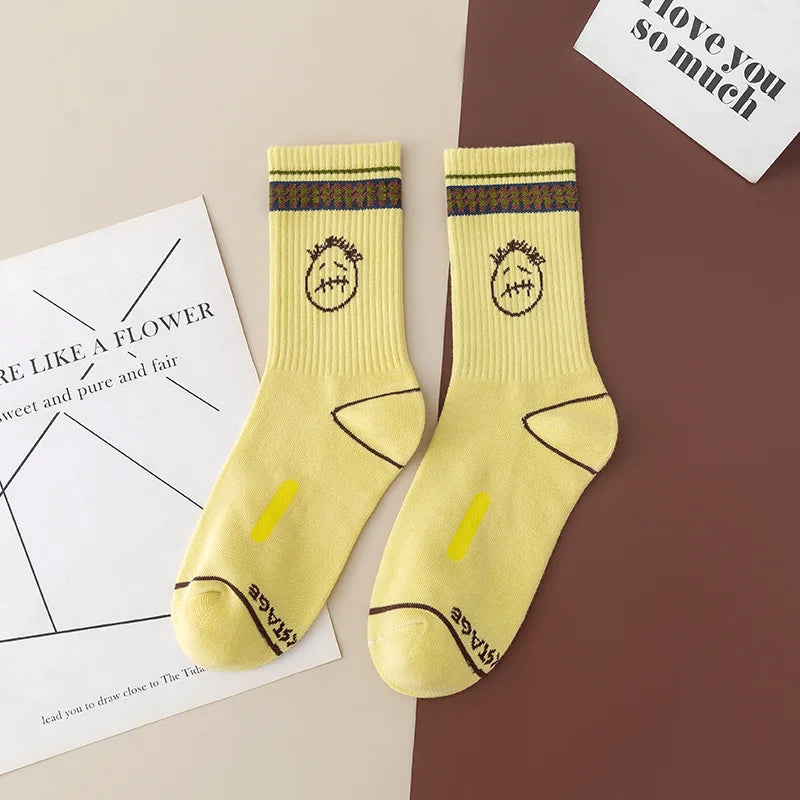 Pale yellow crew socks with dark stitched-face graphics and patterned cuff stripes, flat lay