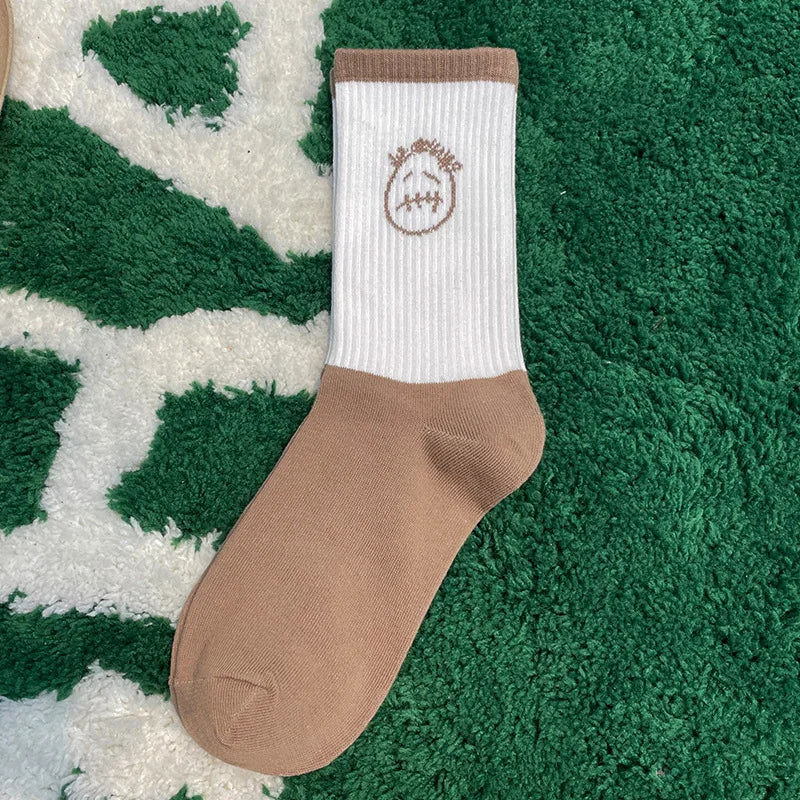 White and tan crew sock with a tan stitched-face graphic, side view on a green rug